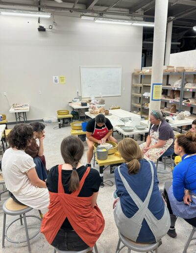 Katie Meili Messersmith demonstrating throwing a bowl on the potters wheel, surrounded by students, intermediate wheel class 1